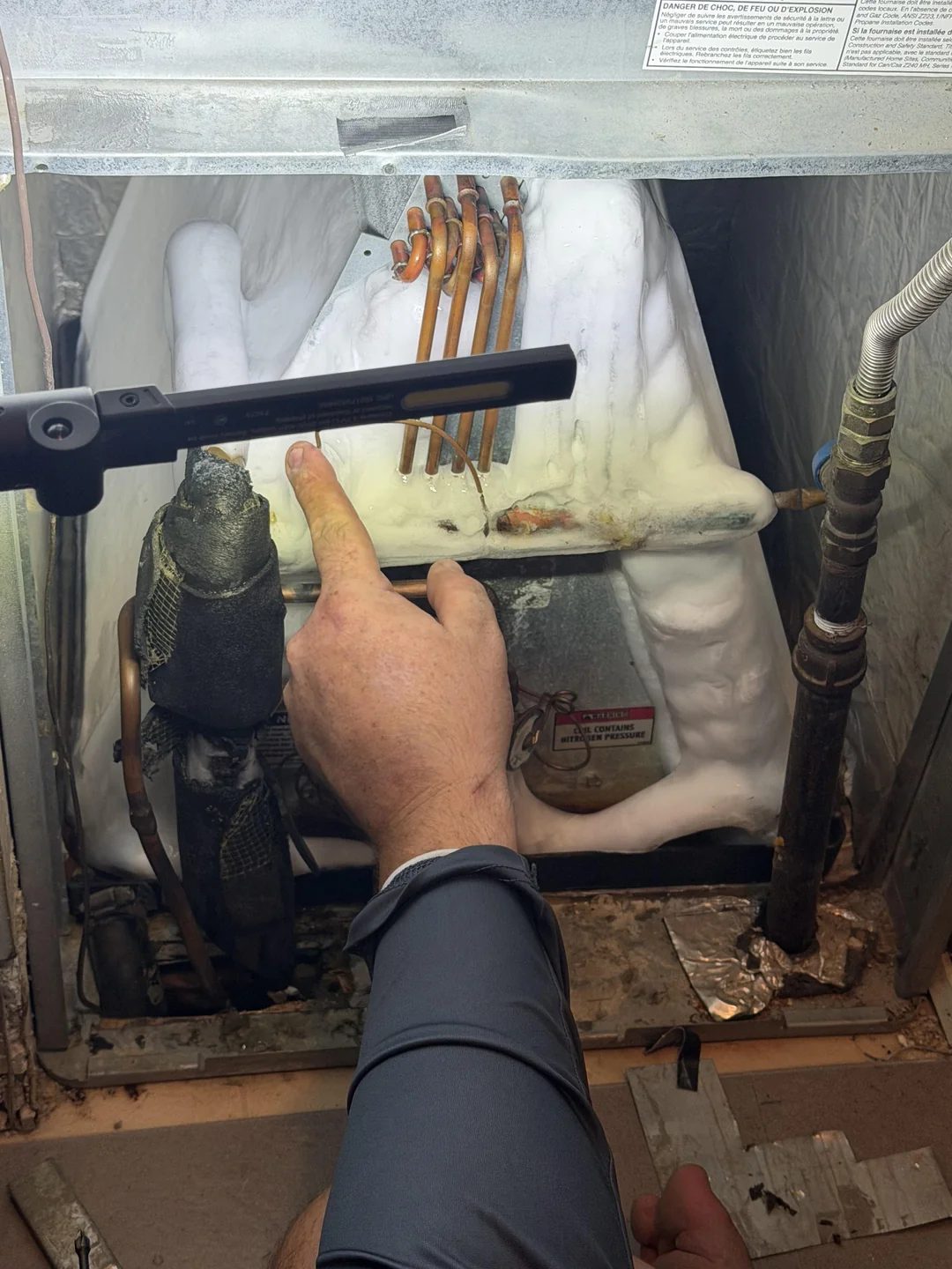 An air conditioning evaporator coil frozen into a solid block of ice inside the furnace cabinet, with a hand pointing at the ice and torn suction line insulation visible beside it